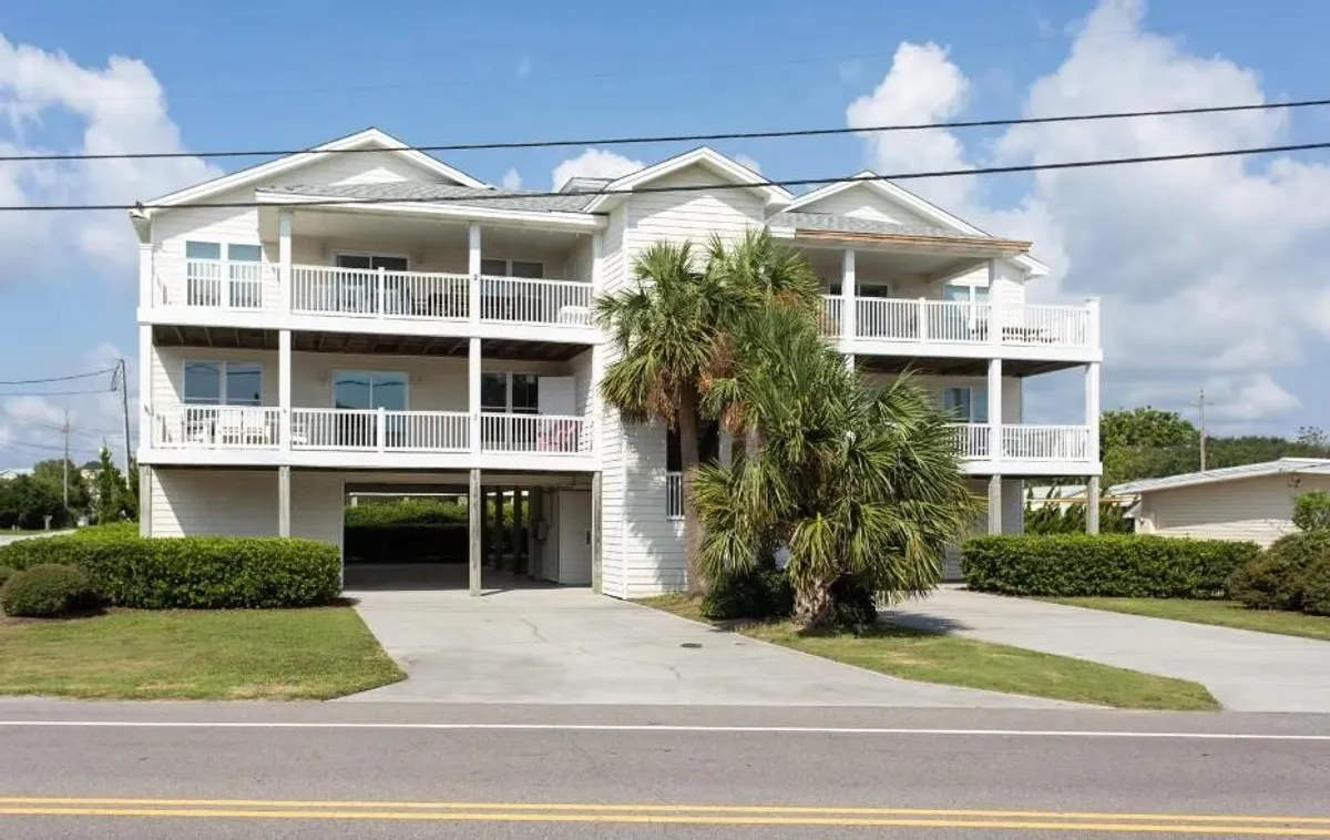 A two-story condo surrounded by palm trees, with spacious balconies ideal for relaxing, dining, or gathering with family. The architecture is open and welcoming, and the setting suggests a tranquil, family-friendly environment just moments from the beach.