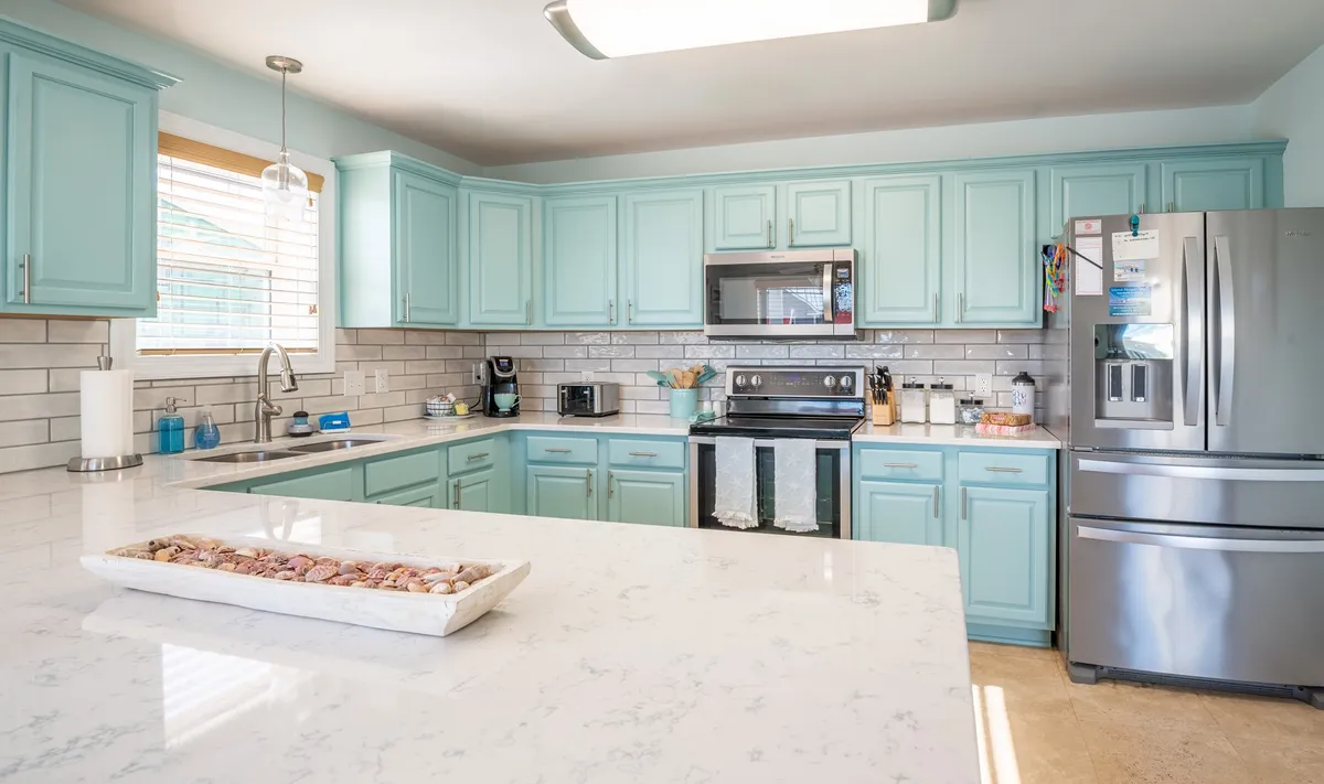 A bright, modern kitchen featuring light blue cabinets, gleaming marble countertops, and high-end stainless steel appliances. Natural light streams in from a window, enhancing the cheerful and inviting atmosphere. The kitchen is designed for both quick snacks and gourmet cooking.