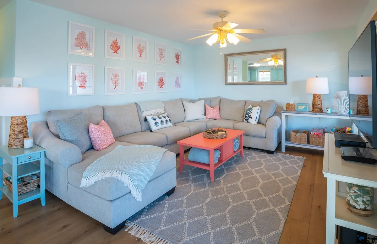 A spacious, modern living room with a large gray sectional sofa centered around a vibrant orange coffee table. The room is decorated with coral-themed wall art and features woven basket decor, exuding a casual, beach-inspired vibe. Ample natural light enhances the inviting atmosphere, making it ideal for gatherings and relaxation.