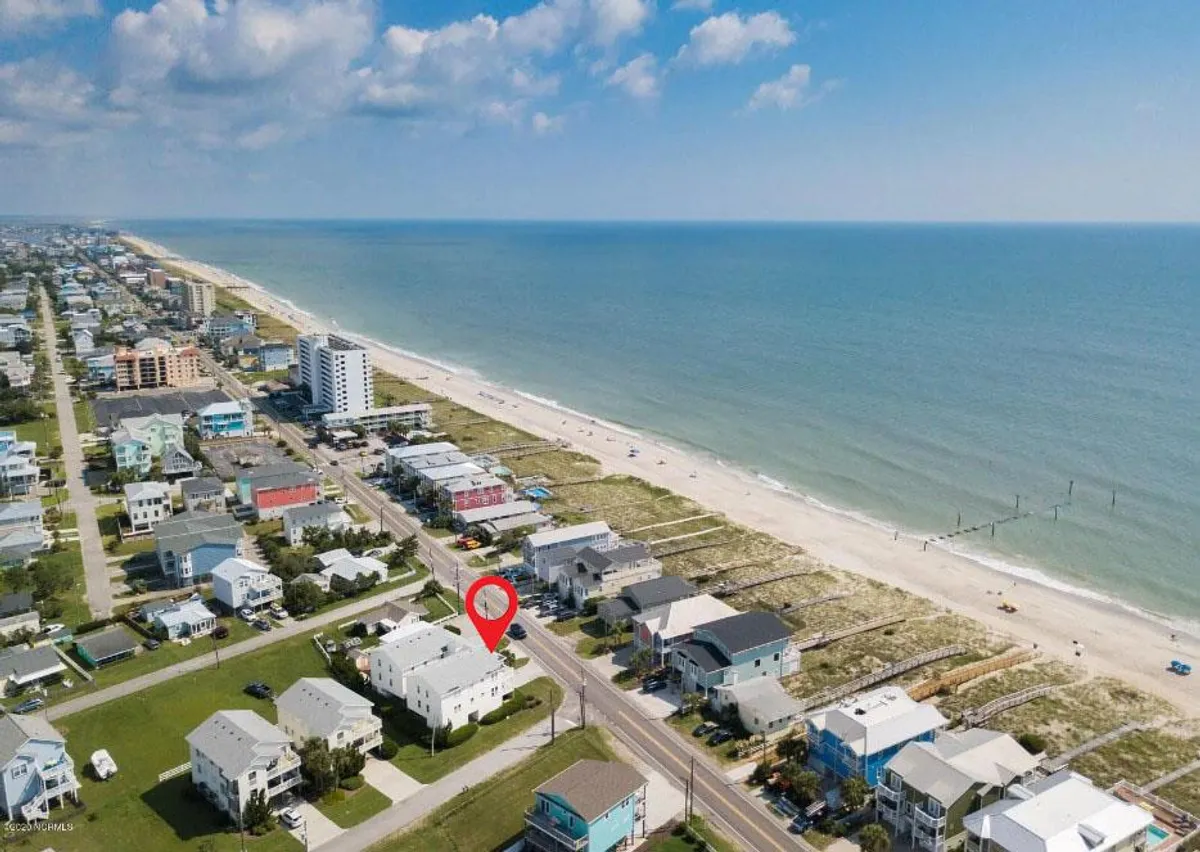A serene view of the beachfront with gentle ocean waves, lined houses, and a wide sandy shore stretching into the distance. The image captures the inviting atmosphere of the coastal location, with soft light suggesting either morning or early evening.