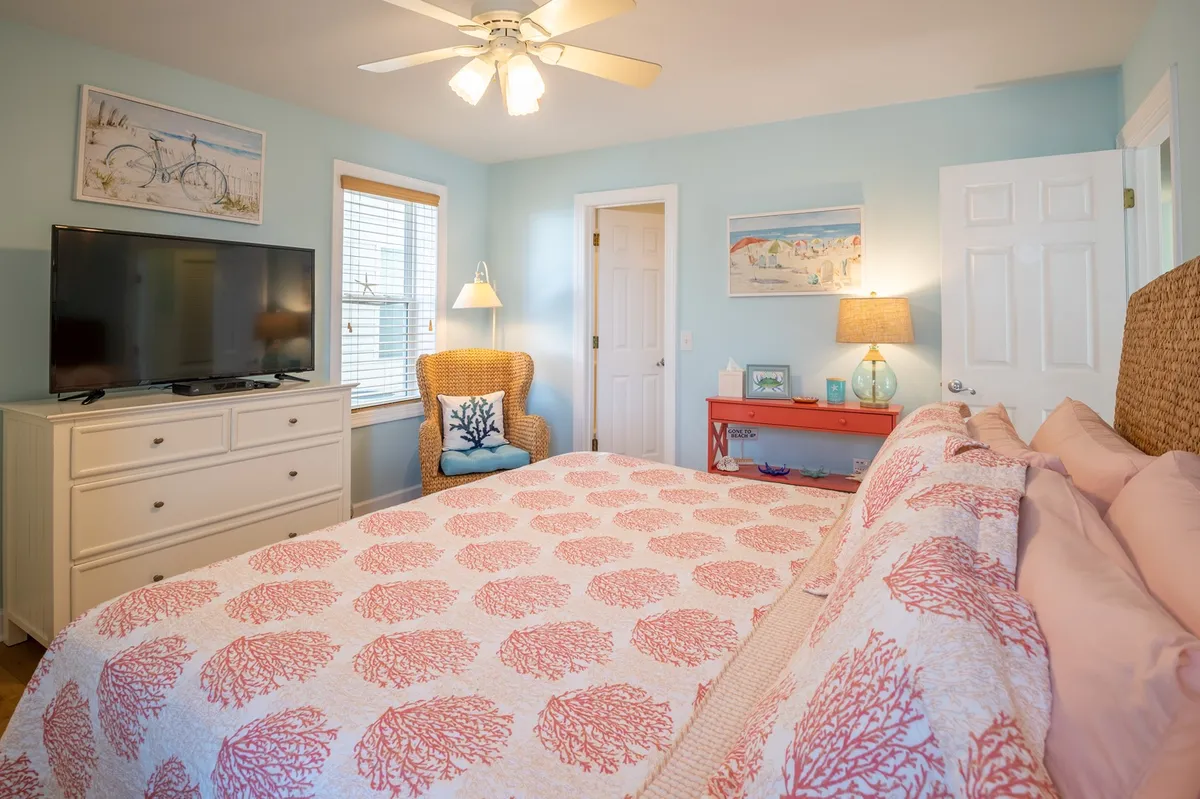 A cheerful bedroom with a coral-patterned quilt on the bed, a wicker chair by the window, and a TV stand. Sunlight streams in, creating a bright and inviting atmosphere perfect for snuggling or enjoying a peaceful evening.