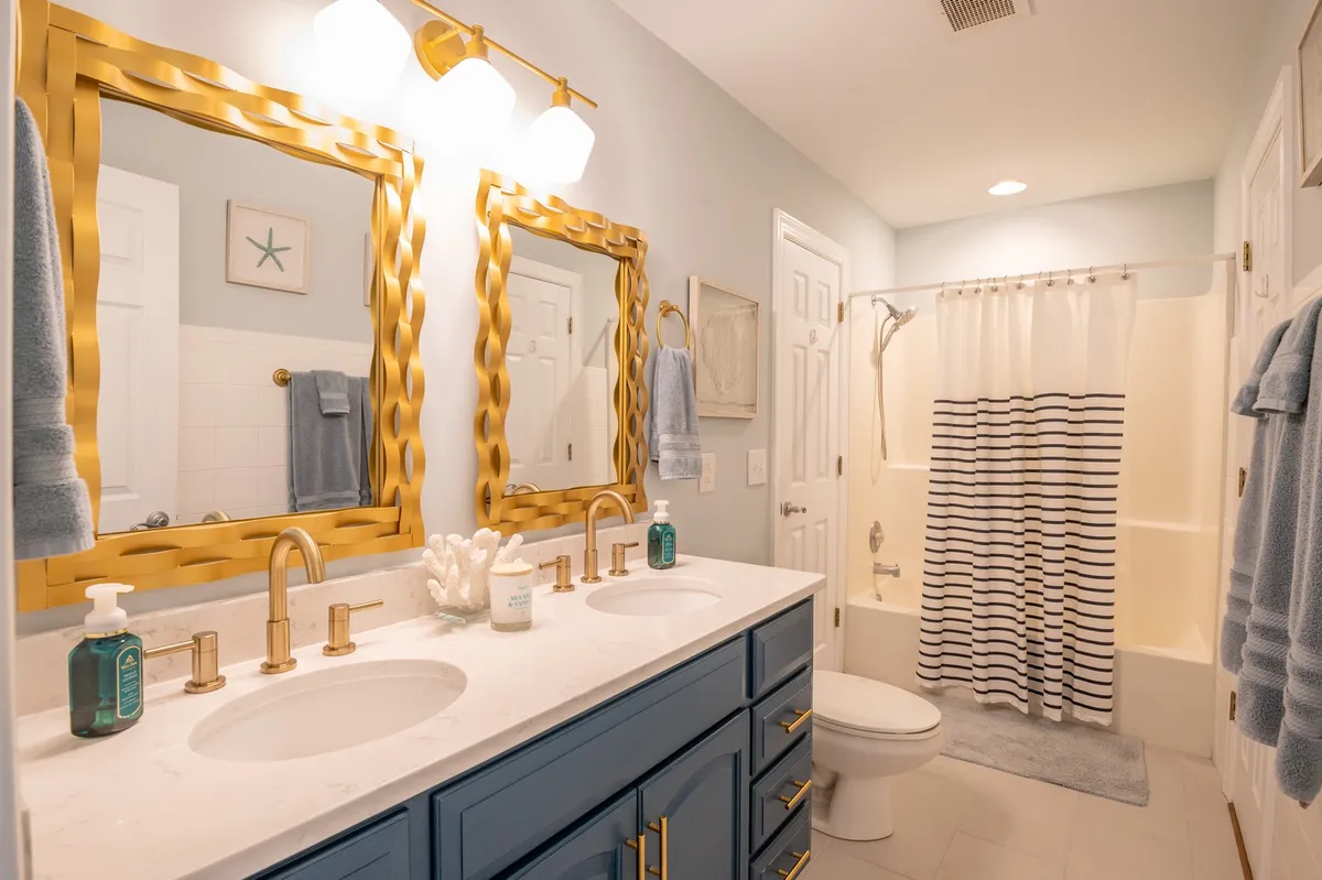 A bright, modern bathroom with dual sinks beneath golden mirrors, a striped shower curtain, and plenty of natural light. The space feels calm and rejuvenating, perfectly suited for morning routines or relaxing baths.