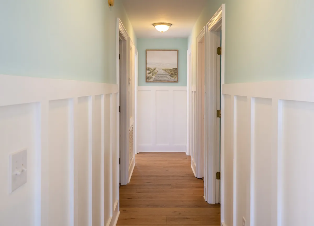A warmly lit interior hallway with natural wood floors, stylish paneling, and framed artwork. The comfortable, inviting design encourages movement through the space, hinting at an open, social layout ideal for groups exploring or gathering within the home.