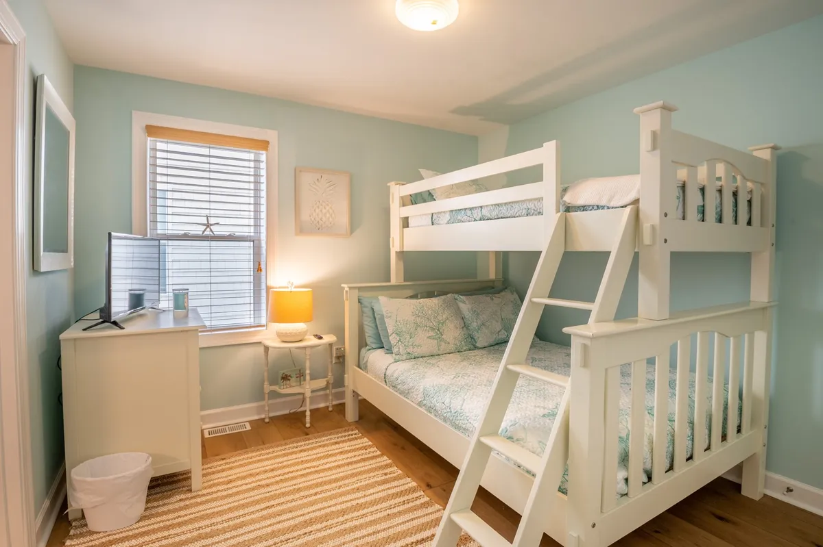 A cheerful bunk room featuring a twin-over-full bunk bed, a sunny window, a desk with lamp for activities or study, and a colorful rug. The space feels playful and practical, appealing to kids or young-at-heart guests seeking a fun environment.