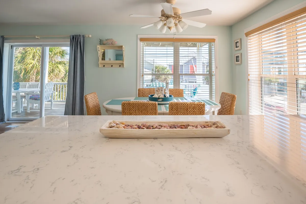 A bright, airy kitchen and dining space featuring a kitchen island with a decorative tray, a table with stylish wicker chairs, and abundant natural light from large windows. The scene feels cheerful and clean, embodying the modern amenities and inviting atmosphere described in the section.