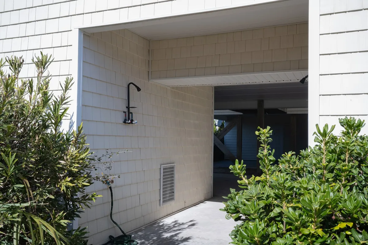 An open exterior entrance flanked by lush greenery and featuring an outdoor shower. The space feels airy and welcoming, with a subtle beach vibe and privacy provided by landscaping.