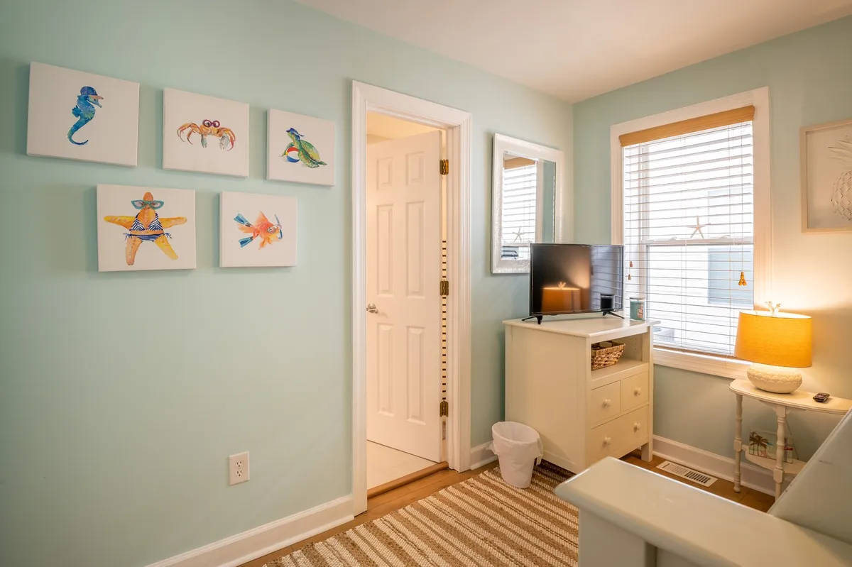 A cozy interior scene featuring a window with blinds letting in natural light, a colorful piece of artwork, a television, and a nightstand with a lamp. The setting feels inviting and intimate, with space for relaxing, enjoying morning coffee, or watching TV together.