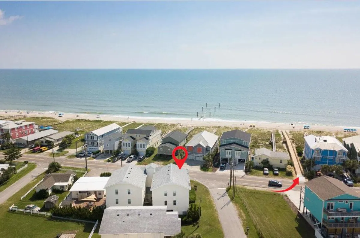 A sweeping view of Kure Beach, with gentle waves, soft sand, and a few beachgoers enjoying the shoreline at sunset. Houses and a pier are visible in the background, offering context to the proximity of the beach to nearby homes. The inviting colors and open space convey the ease of access and the welcoming nature of the beach environment.