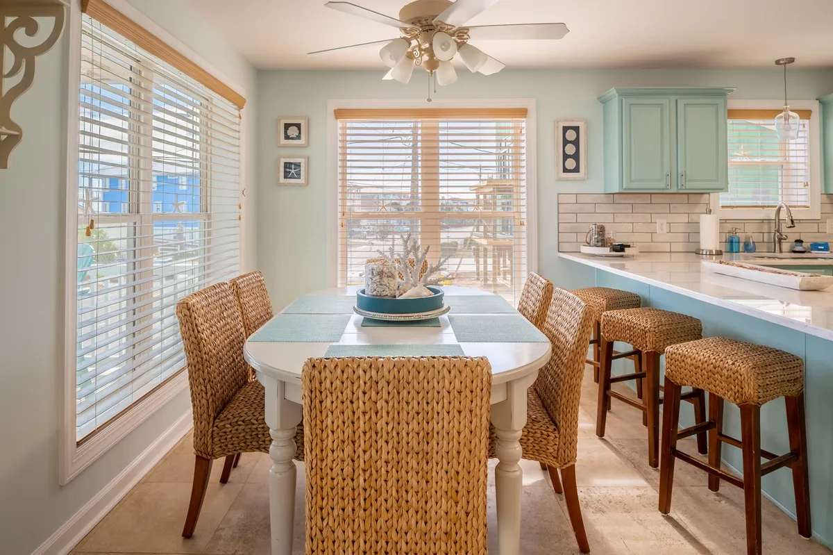 An airy dining area with a solid wood table and woven seagrass chairs, positioned near large windows that offer a glimpse of the ocean. The dining space seamlessly connects to visible kitchen cabinetry, creating a cohesive flow between dining and cooking areas.