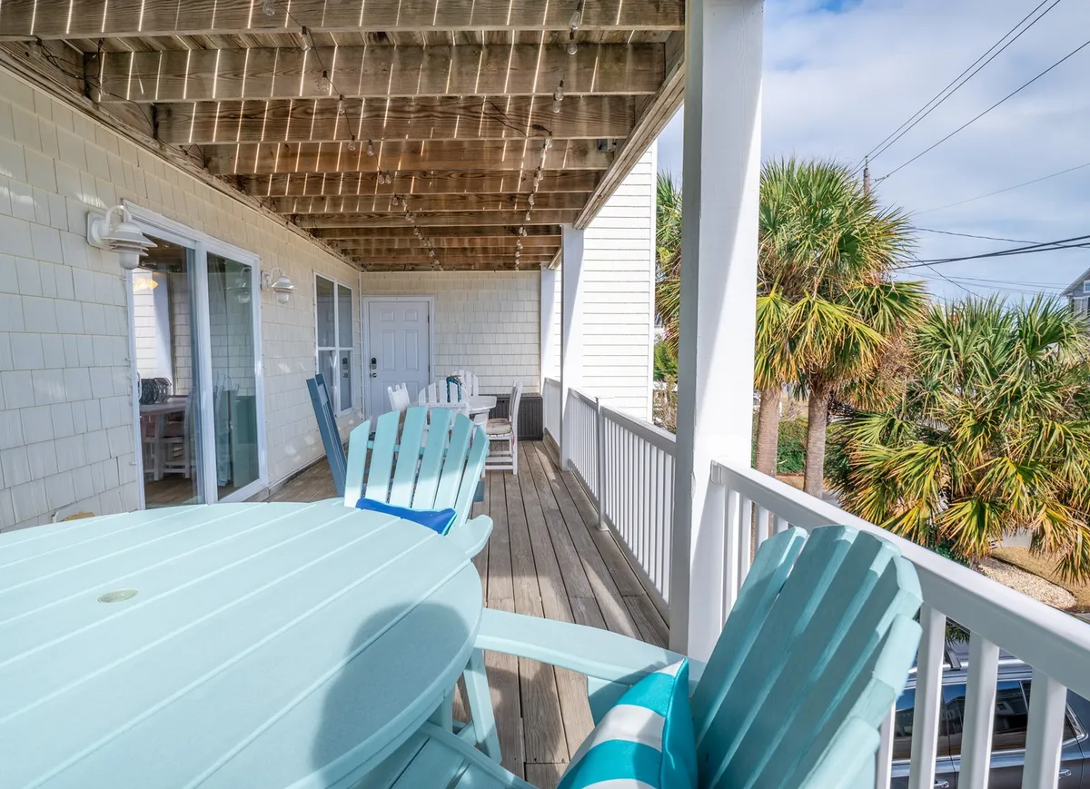 A generous covered porch area decorated with classic blue Adirondack chairs, a wooden table for outdoor dining, and palm trees in the background. Sliding glass doors hint at easy access between interior and exterior spaces, while the porch basks in tranquil natural light.