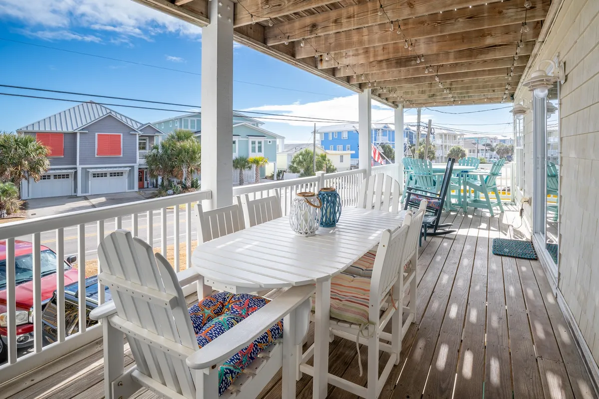 The image shows a cheerful outdoor dining area on the condo's porch, featuring a dining table surrounded by colorful chairs and a decorative vase as the centerpiece. The scene is set for relaxation, with plenty of space for guests to gather for meals, celebrations, or morning coffee while enjoying the fresh coastal air.