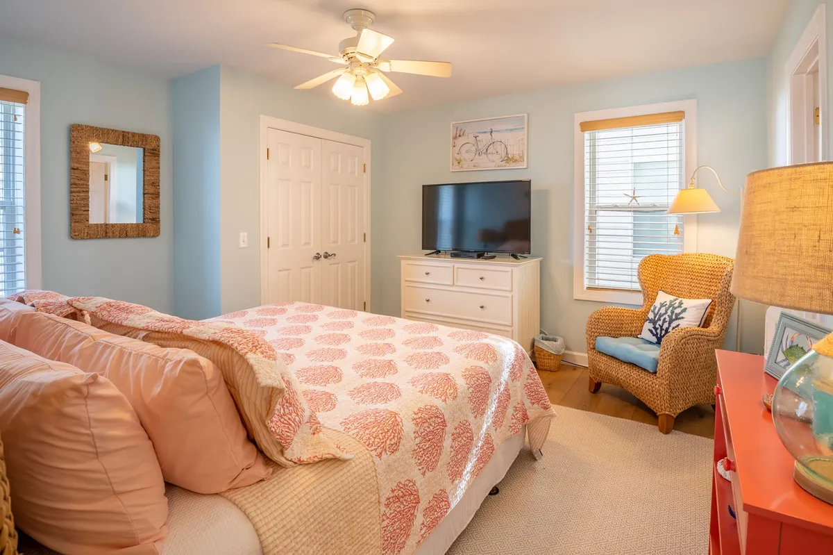 This cheerful bedroom features a bed with a coral-patterned quilt, a wicker chair for reading, and a TV unit. Decorative artwork and natural light create a calm and inviting ambiance, ideal for recharging after outdoor excursions.