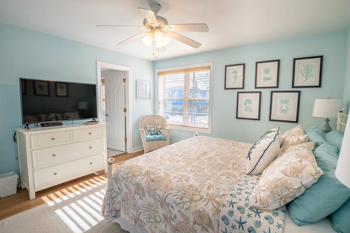 A cozy bedroom scene with a stylishly made bed featuring decorative bedding, a television mounted for nighttime viewing, wall art, a window streaming in natural light, and a wicker chair for reading or relaxing. The mood is calm and inviting, perfect for unwinding after a busy day.