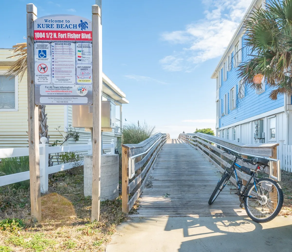 A wooden walkway bordered by manicured landscaping leads towards the beach, accompanied by a friendly welcome sign and a bicycle parked nearby. The path suggests easy, walkable beach access and a family-friendly, relaxed community vibe.